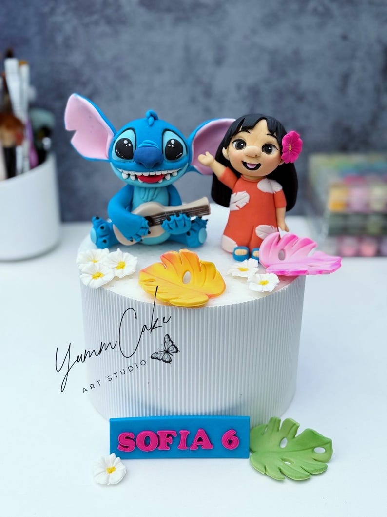 May include: A white cake with a blue and pink nameplate that reads "SOFIA 6". The cake is decorated with figurines of Stitch playing a ukulele and Lilo, along with floral and leaf accents. The cake has the text "Yumm Cake ART STUDIO".