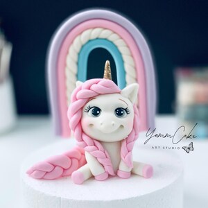 May include: A whimsical cake topper featuring a white unicorn with pink braided hair, a golden horn, and a smiling face. A pastel rainbow arches behind the unicorn. The text "Yumm Cake ART STUDIO" is visible.