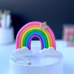 Puede incluir: Un pastel de arcoíris con un arco de arcoíris multicolor, una estrella dorada y nubes blancas. El pastel tiene el texto "Yumm Cake Art Studio" y un logotipo de mariposa. El pastel está sobre una superficie blanca.