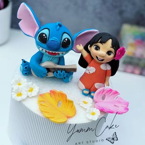 May include: A cake decorated with blue Stitch playing a ukulele and Lilo in an orange dress. The cake is adorned with yellow and pink leaves, white flowers, and the text "Yumm Cake Art Studio."