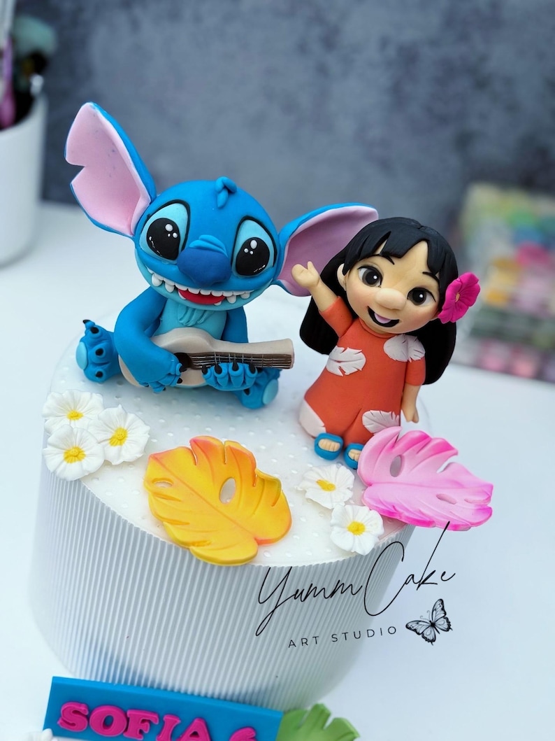 May include: A cake decorated with blue Stitch playing a ukulele and Lilo in an orange dress. The cake is adorned with yellow and pink leaves, white flowers, and the text "Yumm Cake Art Studio."