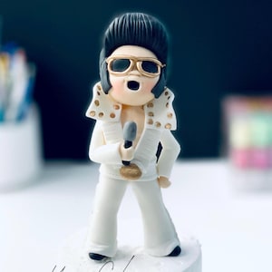 May include: A figurine of a singer in a white jumpsuit with gold detailing, holding a microphone. The figure has black hair, gold-rimmed sunglasses, and an open mouth. The figurine is standing on a white base.