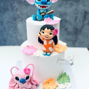 May include: A tiered white cake decorated with cartoon character figurines. The top tier features a blue alien playing a ukulele. The middle tier has a girl waving. The bottom tier has a pink alien. The cake is decorated with flowers and leaves.
