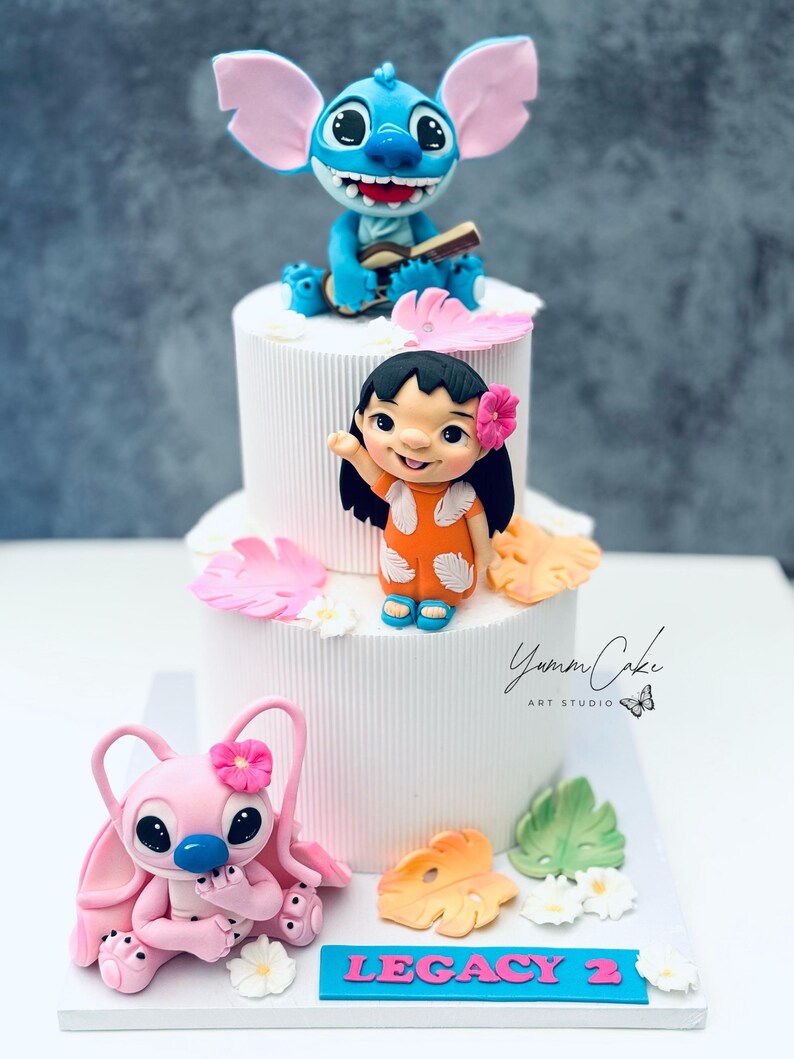 May include: A tiered white cake decorated with cartoon character figurines. The top tier features a blue alien playing a ukulele. The middle tier has a girl waving. The bottom tier has a pink alien. The cake is decorated with flowers and leaves.