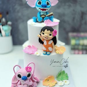 May include: A tiered white cake decorated with blue, pink, and orange character figurines. The cake features Stitch, Lilo, and Angel figures, with floral and leaf accents. The base of the cake has a blue sign that reads "LEGACY 2".