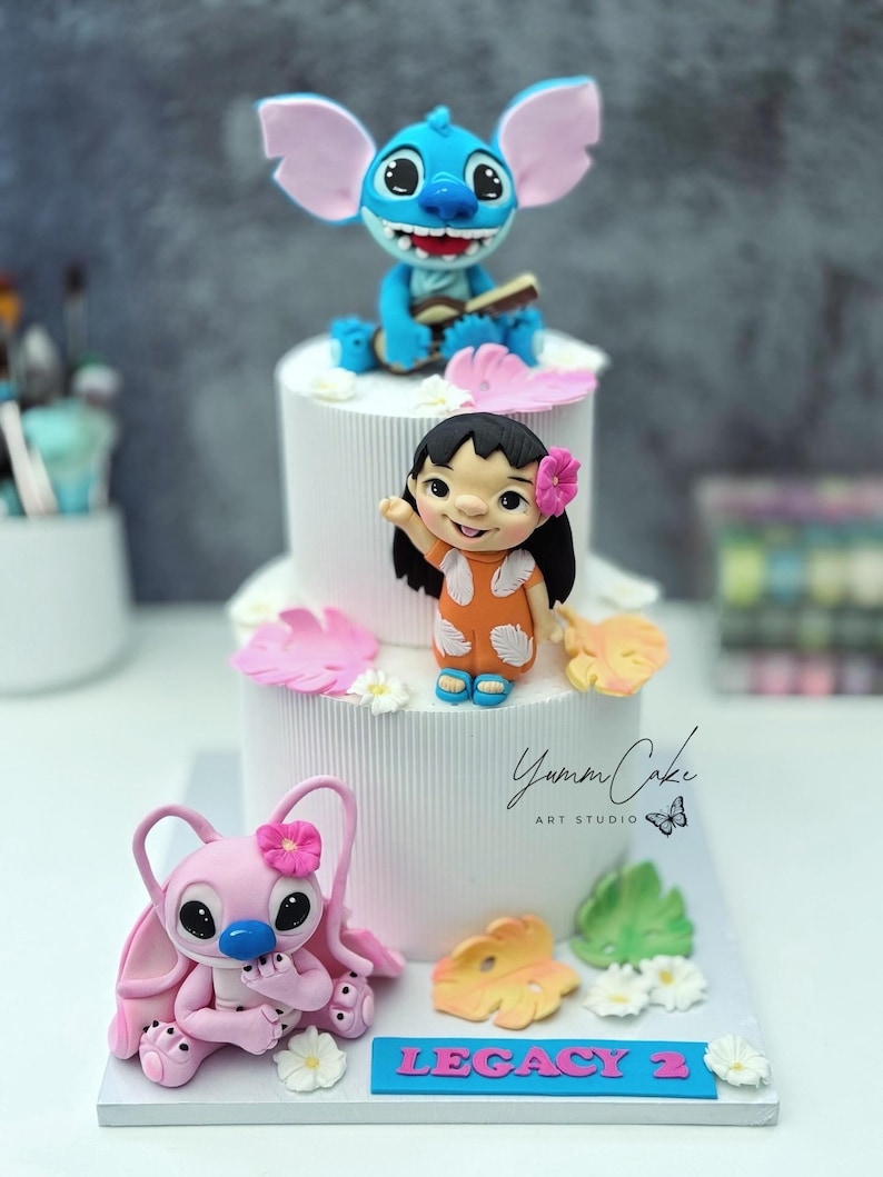 May include: A tiered white cake decorated with blue, pink, and orange character figurines. The cake features Stitch, Lilo, and Angel figures, with floral and leaf accents. The base of the cake has a blue sign that reads "LEGACY 2".