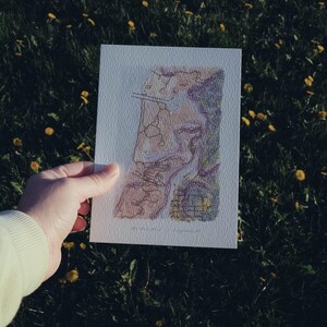 May include: A hand holding a rectangular art print with a map design. The map features shades of brown, purple, and green, with black lines. The print is on textured white paper, set against a backdrop of green grass and yellow flowers.