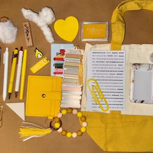May include: A selection of yellow and white stationery and accessories, including a tote bag, journal, pens, and a beaded bracelet. Other items include a keychain, stickers, and a small wallet. The items are arranged on a brown surface.