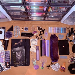 Op de afbeelding: Een assortiment briefpapier en accessoires, waaronder notitieboekjes, pennen, sleutelhangers en stickers, in tinten paars, zwart en zilver. De items zijn gerangschikt op een lichtbruin oppervlak, met een doorzichtige opbergkast op de achtergrond.