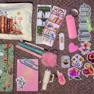 Può includere: Un assortimento di articoli di cancelleria e accessori, tra cui un taccuino con stampa floreale, un diario rosa con un disegno di albero dorato e un astuccio con un'illustrazione di libro e fiori. Sono inclusi anche adesivi, penne e uno scrunchie soffice.