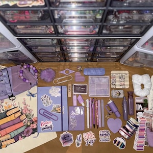 Puede incluir: Una colección de artículos de papelería y accesorios morados, que incluyen cuadernos, bolígrafos, pegatinas y un pequeño bolso. También hay una bolsa de mano con un diseño floral y de libros. Los artículos están dispuestos sobre una superficie marrón, con cajones de almacenamiento en el fondo.