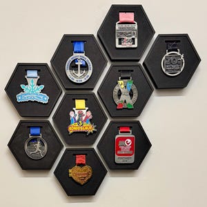Puede incluir: Una colección de medallas coloridas exhibidas en marcos hexagonales negros. Las medallas presentan varios diseños, incluyendo un ancla y el texto "5150 KRAICHGAU". Cada medalla está suspendida por una cinta de diferente color.
