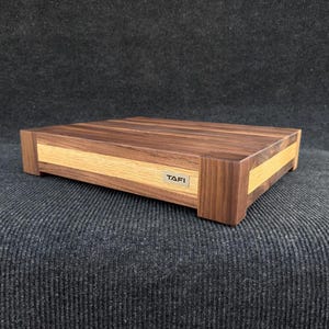 May include: A rectangular wooden box with a light-colored wood accent and a dark wood top. The box has a small metal label that reads "TAFI". The box is on a dark gray surface.