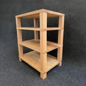 Puede incluir: Una unidad de almacenamiento de madera clara con tres estantes. Los estantes son rectangulares y están soportados por postes verticales en cada esquina. La veta de la madera es visible. La unidad es independiente y se encuentra sobre una superficie gris oscuro.