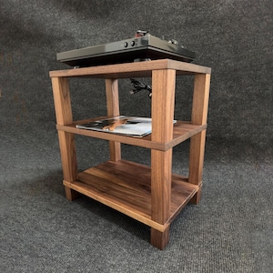 Puede incluir: Un soporte de audio de madera de tres niveles con un tocadiscos en la parte superior. El soporte está hecho de madera marrón oscuro y presenta un diseño rectangular con estantes abiertos. Un álbum de discos es visible en el estante del medio. El soporte está diseñado para equipos de audio.