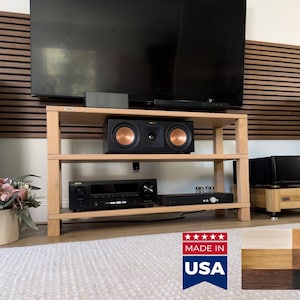 Puede incluir: Un mueble multimedia de madera clara con tres estantes abiertos, que sostiene un altavoz central negro con conos de cobre, un receptor negro y un amplificador negro. Un letrero indica MADE IN USA con seis muestras de color de madera.