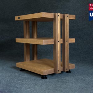 Puede incluir: Un estante de madera para equipos de audio de tres niveles con un acabado de grano de madera marrón claro natural. La estructura consta de tres estantes horizontales sostenidos por cuatro pilares verticales con uniones circulares visibles, apoyados sobre cuatro pies negros.