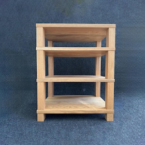 May include: A light brown, three-tiered wooden shelf unit. The shelves are supported by vertical posts at each corner. The unit is made of light-coloured wood and has a simple, minimalist design. The shelf is approximately 61 cm wide, 30 cm deep, and 61 cm tall.