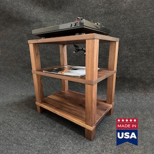 Puede incluir: Un soporte de madera de tres niveles con acabado de veta marrón oscuro, que cuenta con cuatro postes verticales en las esquinas y tres estantes horizontales. Un tocadiscos negro descansa en el estante superior y una revista en el medio.