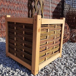 Könnte beinhalten: Ein quadratischer Holzpflanzkübel mit einem geflochtenen Weiden-Design. Der Rahmen besteht aus hellem Holz, und die geflochtenen Seiten weisen natürliche braune und grüne Weidenzweige auf. Dekorative Kugeln sind im Hintergrund.