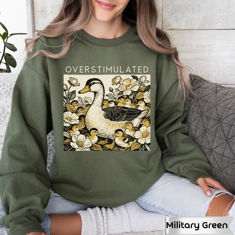 May include: Military green sweatshirt with the word "OVERSTIMULATED" above a graphic of a duck surrounded by ducklings and flowers. The duck is black, white, and tan. The flowers are white and yellow.
