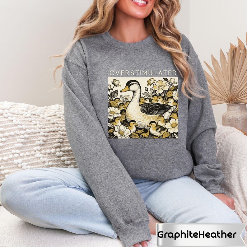 May include: Graphite heather gray sweatshirt with the word "OVERSTIMULATED" above a graphic of a duck and ducklings surrounded by flowers. The sweatshirt is a crew neck style.