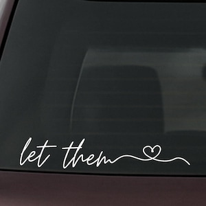 Può includere: Decalcomania in vinile bianco con la scritta "let them" in corsivo, seguita da un simbolo a forma di cuore. Progettata per essere applicata sul finestrino di un'auto o su una superficie simile.