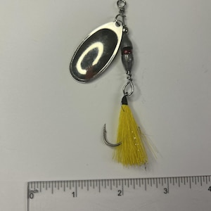 May include: A silver fishing lure with a curved oval blade and a yellow, feathery tail. The lure has a hook and a small red bead. The ruler shows the lure is approximately 2 inches long.