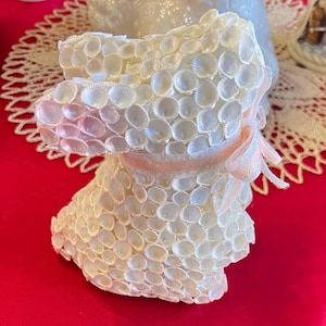 May include: A dog-shaped ornament made from small, white seashells. It features a light pink ribbon tied in a bow. The figurine sits on a red surface, with a lace doily in the background. The overall effect is delicate and decorative.