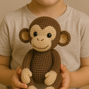 Può includere: Una bambola a forma di scimmia all'uncinetto, marrone con viso, orecchie, mani e piedi beige. La scimmia ha occhi neri e un sorriso cucito. La bambola è tenuta da una persona.
