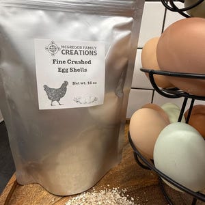 May include: A silver pouch labeled "McGregor Family Creations Fine Crushed Egg Shells" with a hen illustration. The net weight is 16 oz. Crushed eggshells are scattered on a wooden surface. A wire egg holder contains various colored eggs.