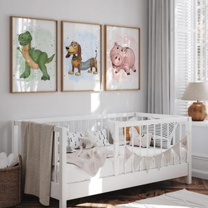 May include: Three framed art prints featuring cartoon characters: a green dinosaur, a brown dachshund with a spring body, and a pink pig. The prints are displayed above a white crib with a beige blanket and decorative pillows.