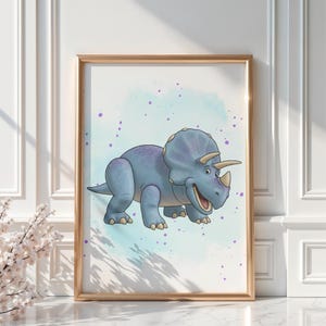 May include: A framed print featuring a cartoon Triceratops dinosaur in shades of blue and purple. The dinosaur has three horns and a friendly expression, set against a watercolor-style background with purple speckles. The frame is gold.