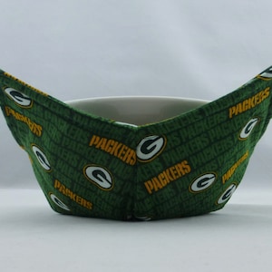May include: Green and gold bowl cozy with the word "Packers" repeated in a pattern. The cozy is designed to hold a bowl and keep it warm.
