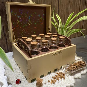 Peut inclure: Une boîte en bois beige avec un couvercle à charnière, contenant de petits bocaux en verre avec des bouchons en liège. La boîte est ouverte, révélant les bocaux disposés en rangées. Un petit bocal en verre est renversé.