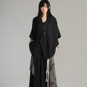 May include: Black kimono-style top with a wrap design and short sleeves. The top is paired with a long black skirt and a decorative necklace. A cream-colored tie with a decorative pendant hangs from the waist. The outfit has a minimalist, modern aesthetic.