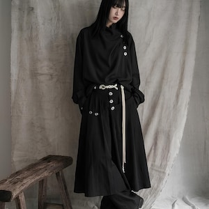 Könnte beinhalten: Ein schwarzes Langarmshirt mit asymmetrischer Knopfleiste am Kragen, kombiniert mit einem schwarzen Midirock mit durchgehender Knopfleiste, einer silberfarbenen Gürtelschnalle und einer langen weißen Kordel an der Taille.