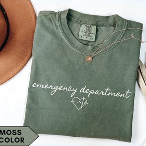 Puede incluir: Una camiseta verde musgo con las palabras "emergency department" en escritura blanca, junto con un corazón y un gráfico de línea de ECG. También se ven un collar dorado y una planta seca.