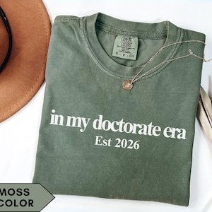 Puede incluir: Camiseta verde musgo con el texto "in my doctorate era Est 2026" en blanco. Un collar dorado con un colgante descansa sobre la camiseta. Un sombrero marrón y una planta seca están en el fondo.