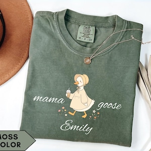 May include: Moss green t-shirt with a cartoon goose wearing a bonnet and holding a drink. The text "mama goose Emily" is printed on the shirt. A gold necklace is draped across the shirt.