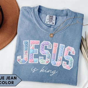 Puede incluir: Camiseta azul vaquero con la palabra "JESUS" en letras de patchwork. La camiseta también tiene la frase "is king". Un collar de oro con un colgante está sobre la camiseta.