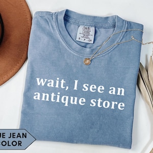 Puede incluir: Una camiseta de color azul vaquero con el texto "wait, I see an antique store" en blanco. También se ve un collar dorado con un colgante y un sombrero marrón.