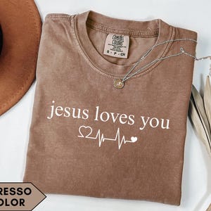 Puede incluir: Camiseta color espresso con el texto blanco « jesus loves you », junto con un corazón y un gráfico de latido cardíaco. Un collar plateado con un colgante descansa sobre la camiseta. La camiseta está doblada y se muestra con un sombrero marrón y una planta seca.