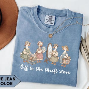 Puede incluir: Camiseta azul vaquero con una ilustración caprichosa de gansos vestidos con atuendos vintage, con carritos de compras y espejos. El texto "Off to the thrift store" está impreso debajo. También se ve un collar dorado y un sombrero marrón.