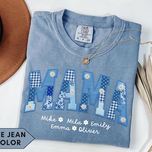 Puede incluir: Camiseta azul vaquero con la palabra "MAMA" en letras de patchwork. Las letras son de varios tonos de azul con estampados florales y de cuadros. Los nombres están bordados debajo. Un collar dorado y un sombrero marrón también están en la imagen.