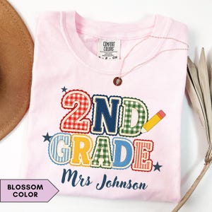 Puede incluir: Una camiseta rosa claro con el texto 2ND GRADE en letras coloridas, estampadas y con bordes festoneados, junto a un gráfico de un lápiz amarillo. Debajo del texto, el nombre Mrs Johnson está impreso en letra cursiva azul oscuro. Etiqueta Comfort Colors.