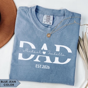 Puede incluir: Camiseta de color azul vaquero con la palabra "DAD" en letras blancas grandes. Debajo de la palabra "DAD" están los nombres "Michael" e "Isabella" con un símbolo de corazón entre ellos. El texto "EST.2026" está debajo de los nombres. Un collar dorado está sobre la camiseta.