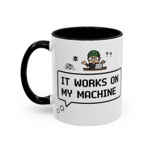 Może przedstawiać: Biały ceramiczny kubek z czarnym wnętrzem i uchwytem. Kubek przedstawia pikselową grafikę osoby z laptopem i tekstem "IT WORKS ON MY MACHINE" w dymku.