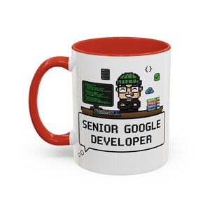 May include: White ceramic mug with a red interior and handle. The mug features a pixelated graphic of a person with glasses and a green beanie, and the words "SENIOR GOOGLE DEVELOPER" in a speech bubble. A computer screen and keyboard are also visible.