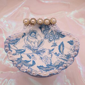 Peut inclure: Un vide-poches en forme de coquillage avec un motif floral bleu sur fond blanc. Le bord est orné de petites perles et le bord supérieur comporte cinq ornements de perles et de strass. Le plat a une finition nacrée.